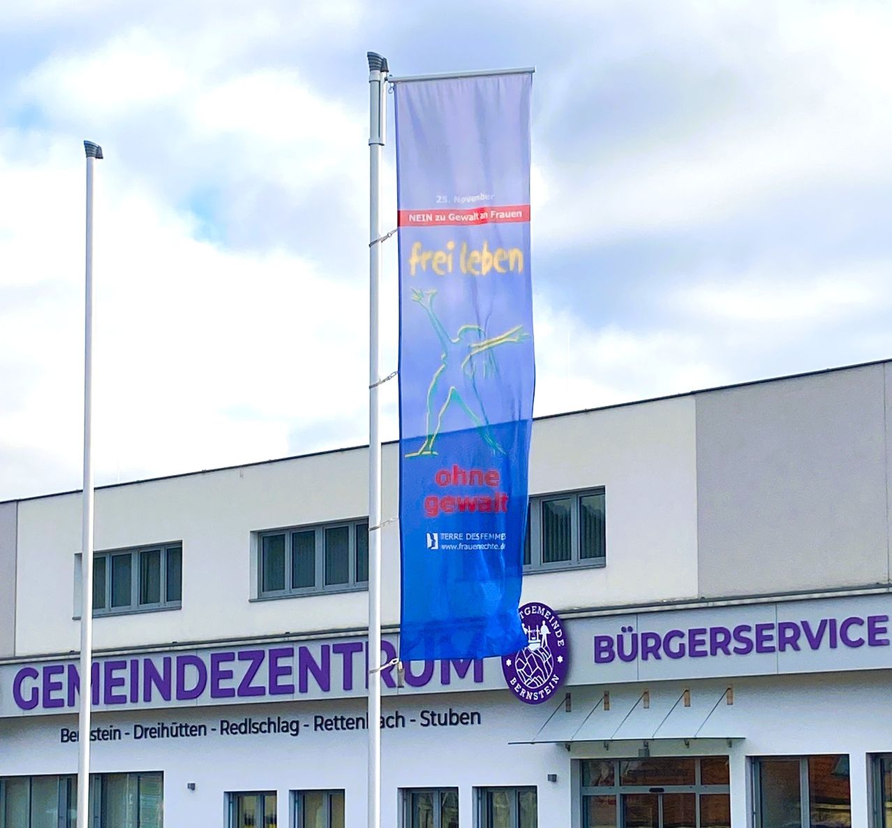 Ein Gebäude mit mehreren Fenstern zeigt ein Banner mit blauem Hintergrund und einem lila Logo. Das Banner enthält die Aufschrift 'frei leben' und 'ohne Gewalt'. Das Gebäude heißt Burgerservice und befindet sich in Stein-Dreihutten.