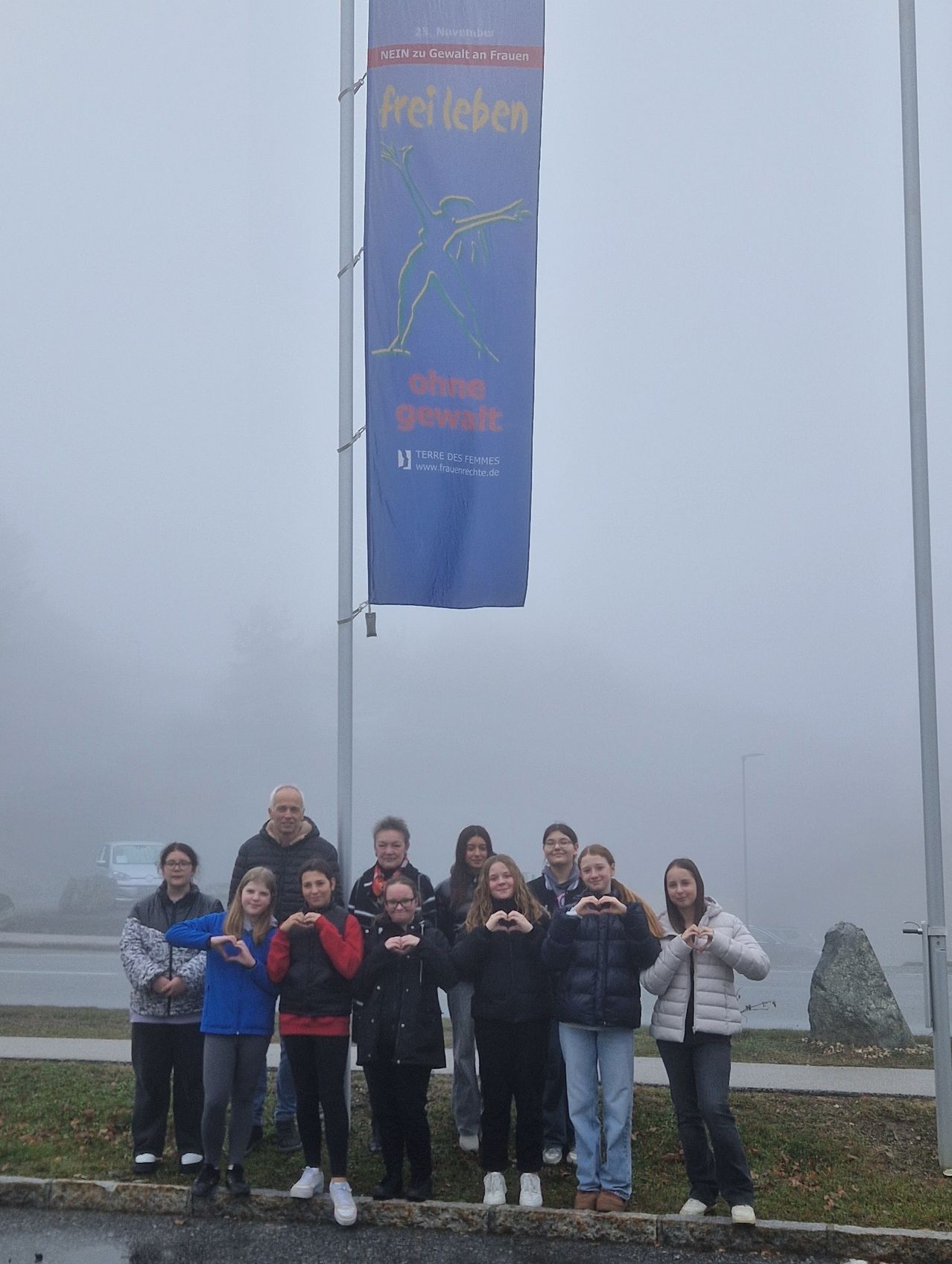 Eine Gruppe von Menschen in Jacken posiert für ein Foto vor einem Pfosten mit einem blauen Banner. Auf dem Banner stehen die Worte 'ohne Gewalt' geschrieben.