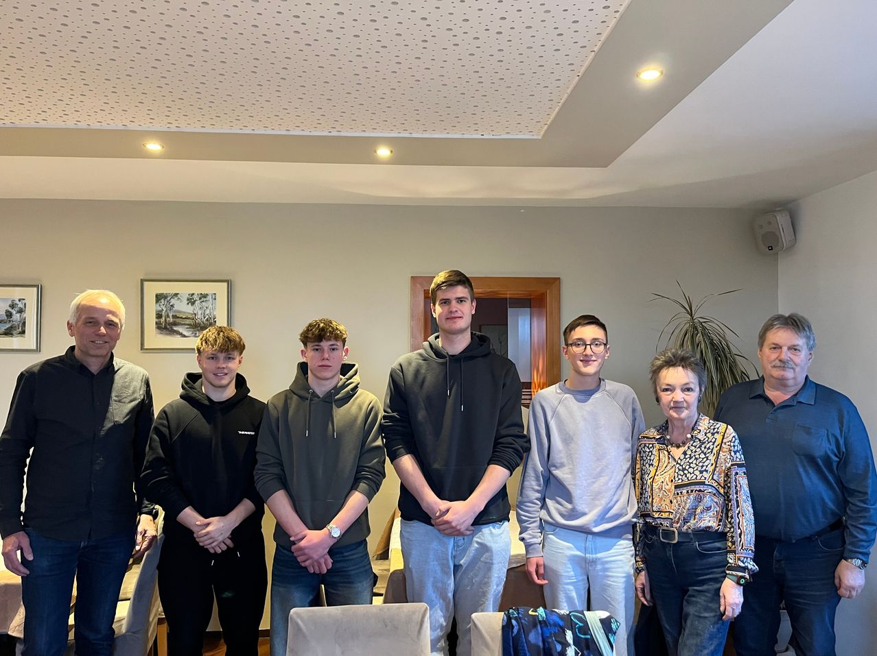 Eine Gruppe von Menschen, darunter mehrere junge Männer und eine ältere Frau, posiert für ein Foto in einem Raum mit weißen Wänden und einer Decke mit quadratischen Lichtern.