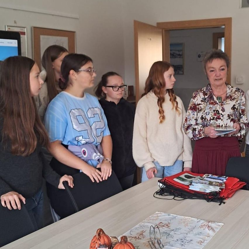 A group of people are standing in a room. An elderly woman is standing near the table. A girl is holding a stuffed toy in her hands. A few pumpkins are on the table.