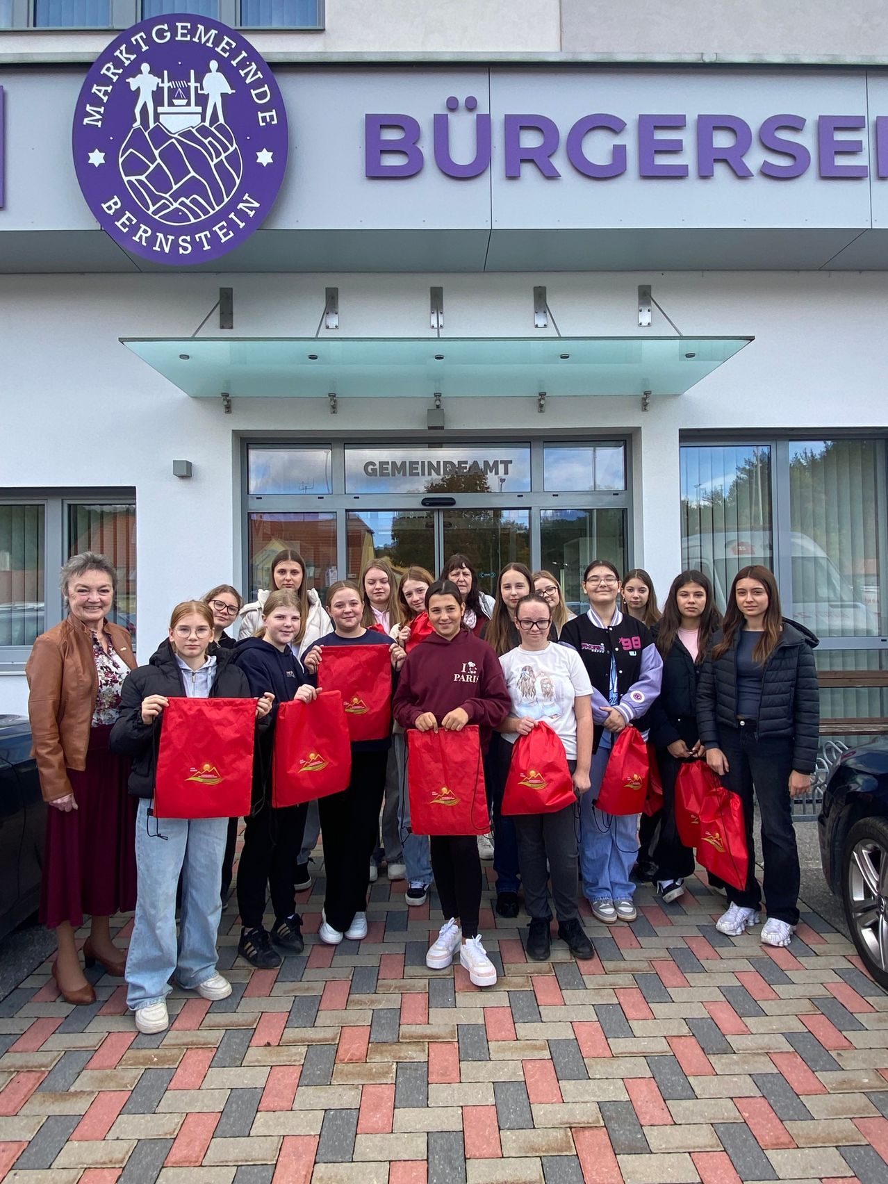 A group of students with red bags stands in front of a building with the sign Burgermeister Bernstein.