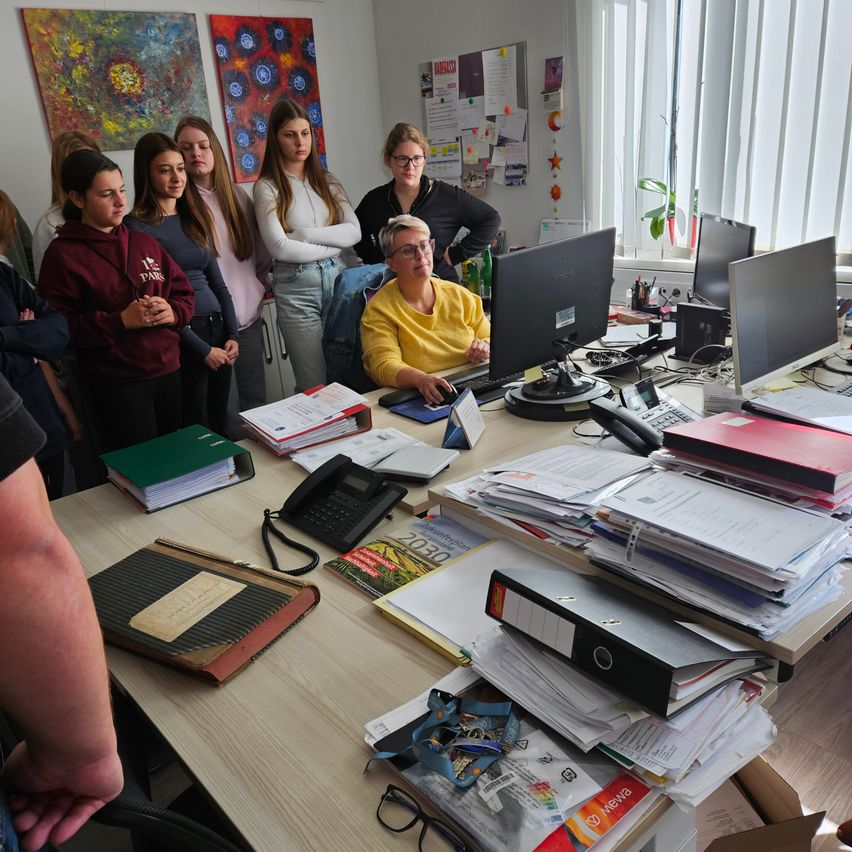 A woman sits at a desk with a computer and various documents, while a group of students stand behind her. Papers, books, and a telephone are on the desk. Art pieces hang on the wall, and a potted plant is by the window.