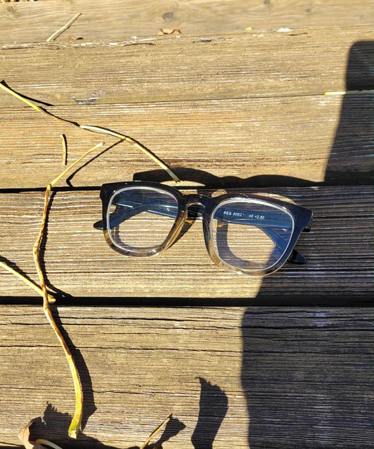 A pair of glasses with clear lenses and a black frame, labeled PEG 60C2 45 +2.50, lies on a wooden surface.