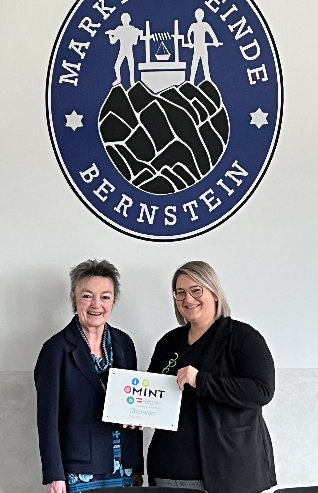 Two women stand smiling in front of a logo that reads Bernstein. The woman on the left holds a white card with the text Mint and some icons. They are both wearing glasses and dark coats.