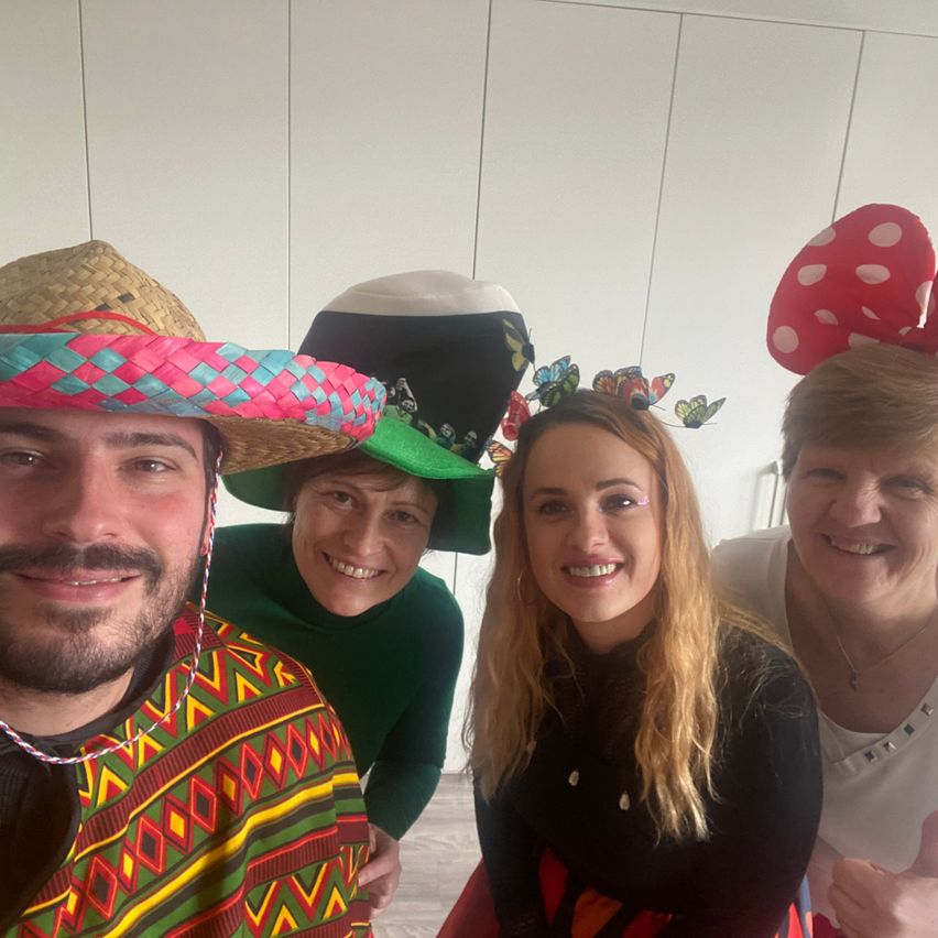 Four individuals, wearing festive hats, are posing for a selfie. A man with a colorful hat, a woman with a butterfly hat, and two others with cheerful expressions. They are in a room with white walls.