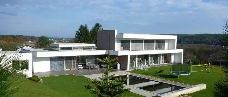 Ein weisses modernes Haus mit einem Swimmingpool und einer Trampolin in der Mitte eines grasigen Feldes.