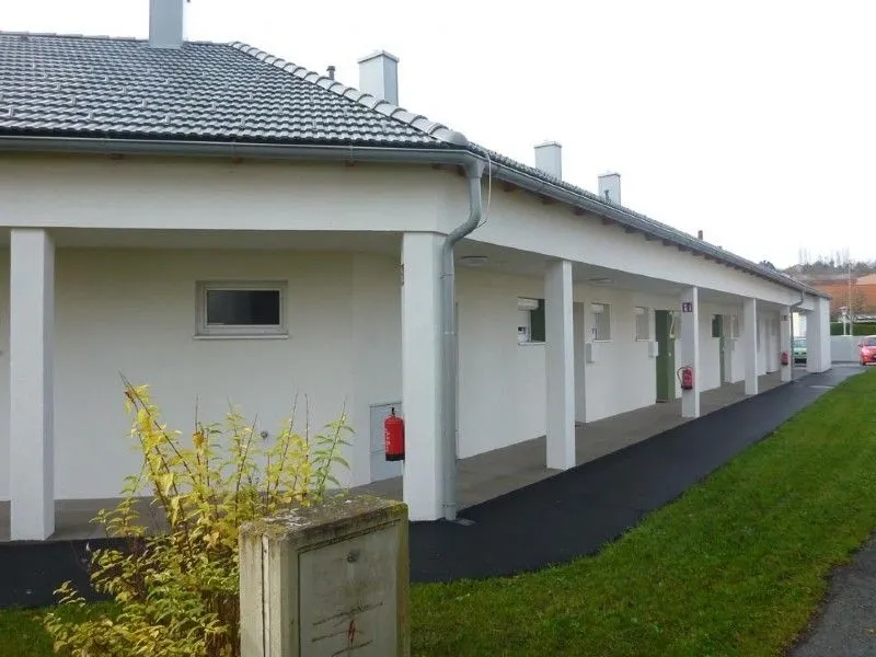 Ein langes weißes Gebäude mit grauem Dach, mehreren Fenstern und zwei roten Feuerlöschern an der Wand.