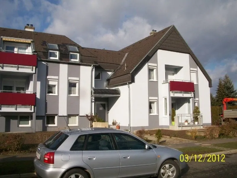 Ein silberner Wagen ist vor einem modernen weißen Mehrfamilienhaus mit roten Markisen geparkt. Der Himmel ist bewölkt.