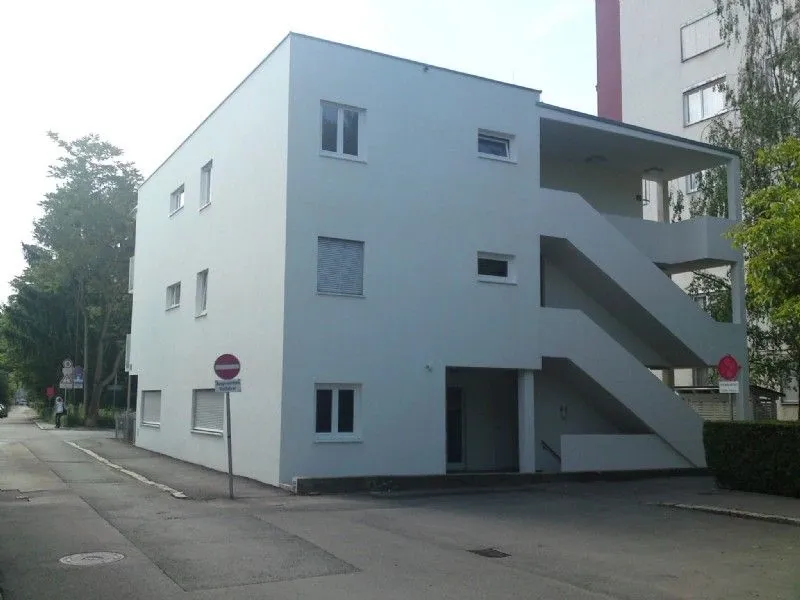 Ein weißes Mehrfamilienhaus mit mehreren Fenstern und einer Außentreppe. Ein rotes und weißes Schild steht am Bürgersteig.