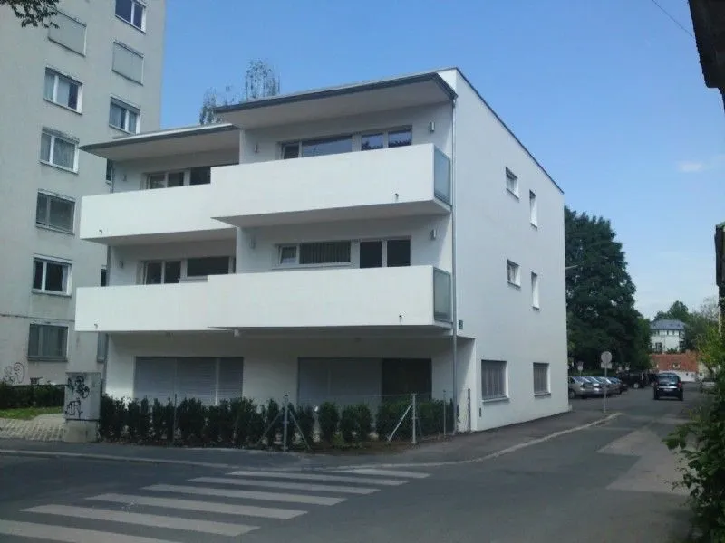 Ein weisses Mehrfamilienhaus mit mehreren Balkonen und Fenstern steht an der Straßenecke. Ein Auto ist am Straßenrand geparkt.
