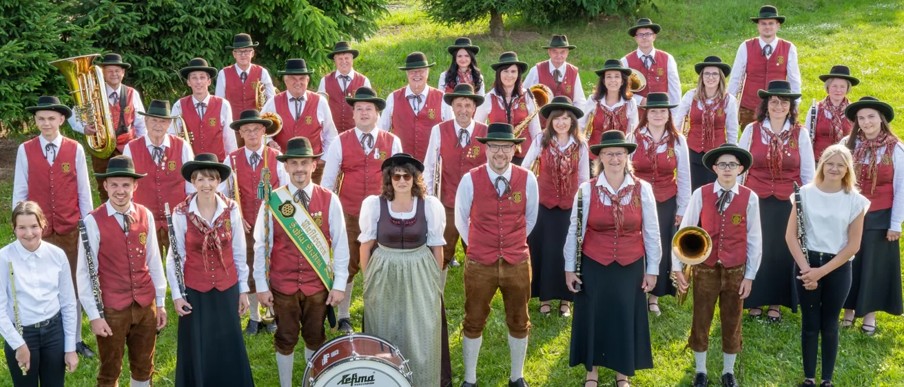 Eine Gruppe von Menschen in traditioneller Kleidung steht auf einem Rasen. Sie sind Musiker und halten verschiedene Instrumente. Ein Schlagzeug und eine Trompete liegen vor ihnen auf dem Boden.