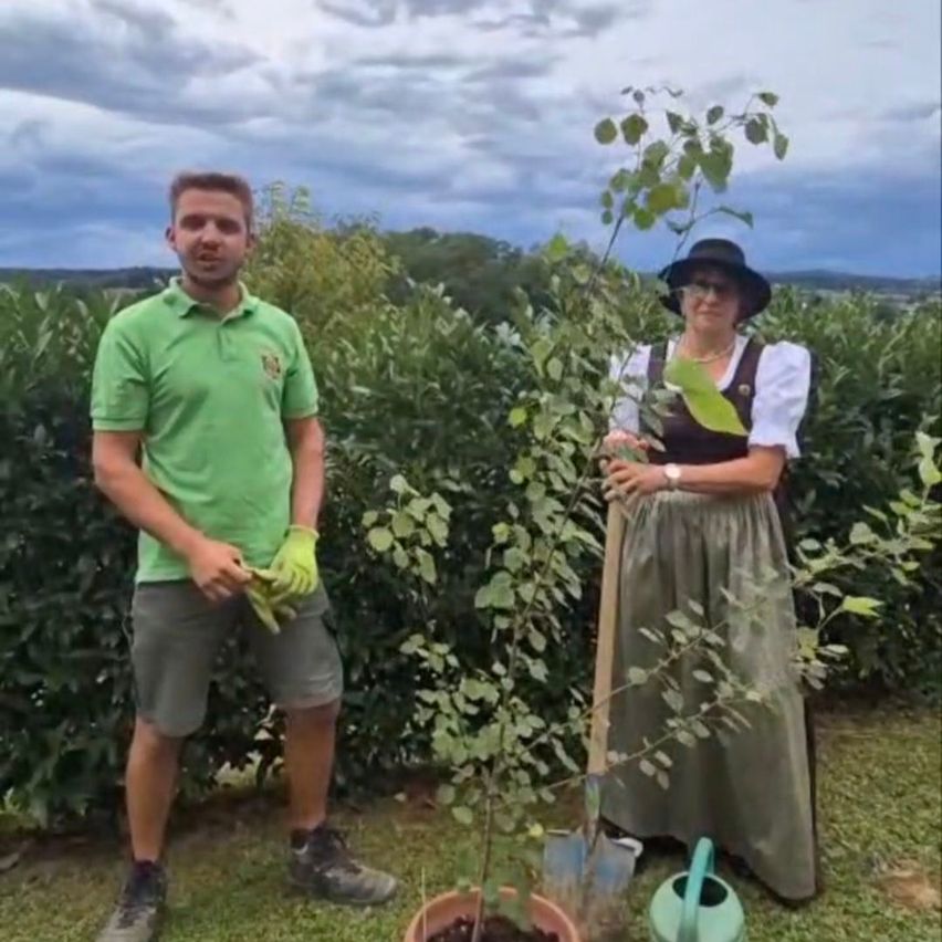 Ein Mann und eine Frau stehen in einem Garten mit einem kleinen Baum, beide tragen grüne Handschuhe und halten Gartenwerkzeuge in der Hand.