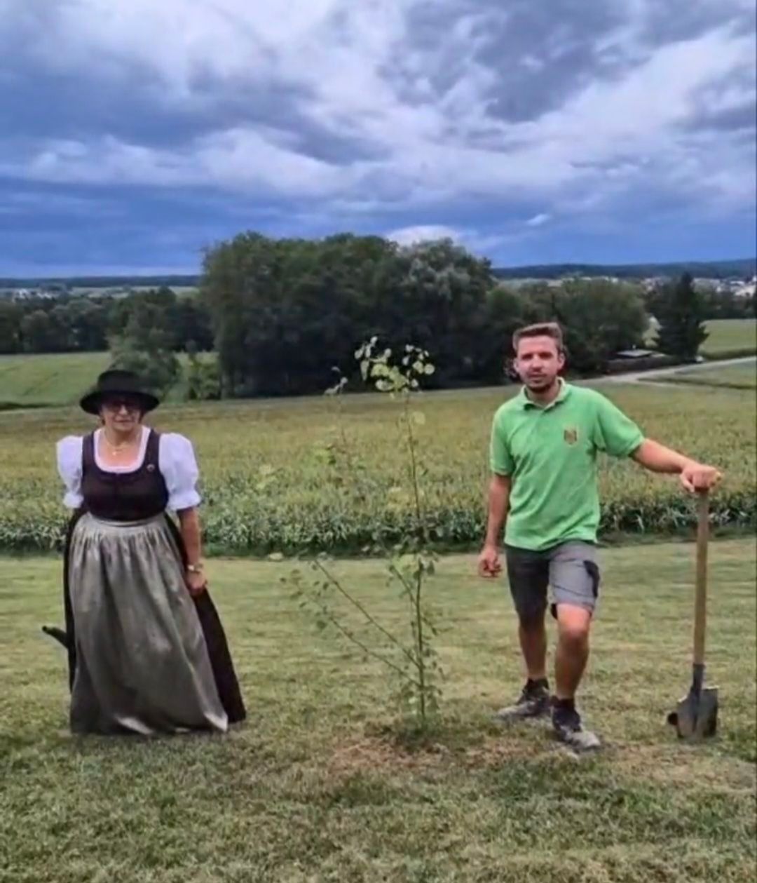 Ein Mann in einem grünen T-Shirt und Shorts hält eine Schaufel und steht neben einer Frau in traditioneller Kleidung auf einem grasbewachsenen Feld.