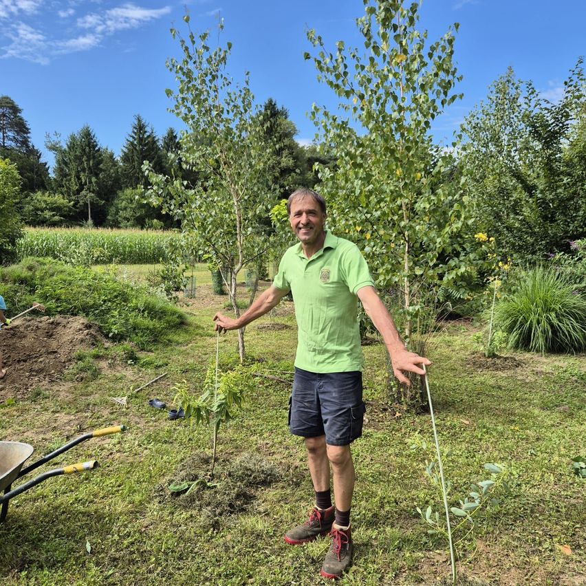 Ein Mann in einem grünen Shirt und blauen Shorts steht in einem Garten mit einem Schubkarren und Pflanzen. Er hält einen Stock und lächelt für die Kamera.