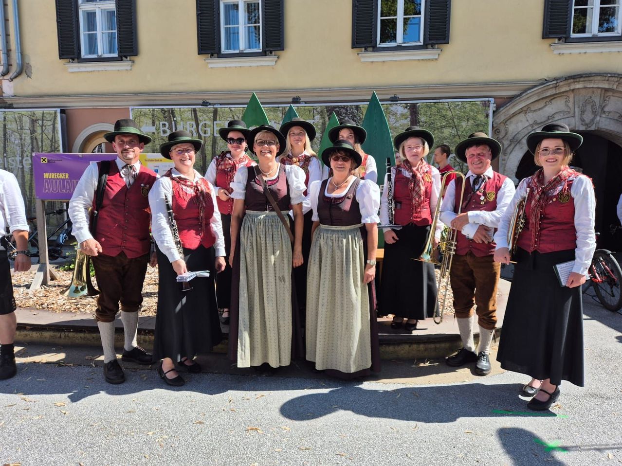 Eine Gruppe von Menschen in traditioneller Kleidung posiert für ein Foto vor einem Gebäude mit Fenstern und einem Banner. Sie halten Musikinstrumente wie Trompeten und Klarinetten.