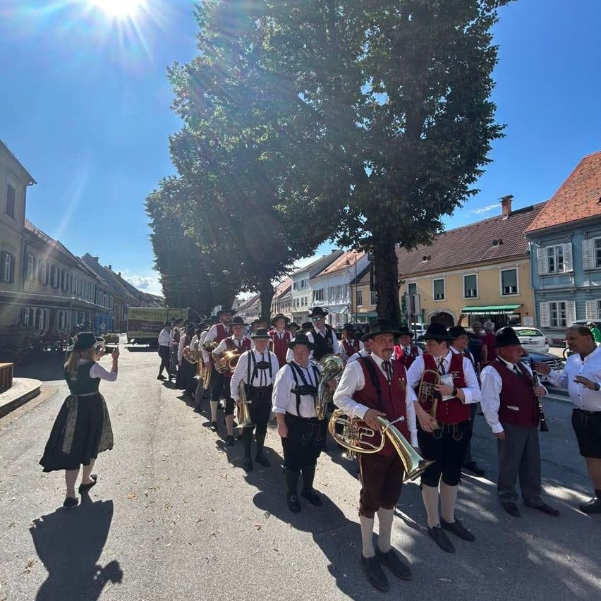 Ein Blasorchester in traditioneller deutscher Kleidung tritt auf einem Stadtplatz bei sonnigem Wetter auf.
