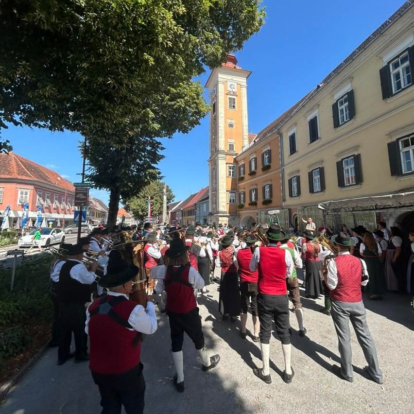 Eine Blaskapelle in traditioneller bayerischer Kleidung tritt auf dem Stadtplatz auf, mit Gebäuden, Bäumen und einem Uhrturm im Hintergrund.