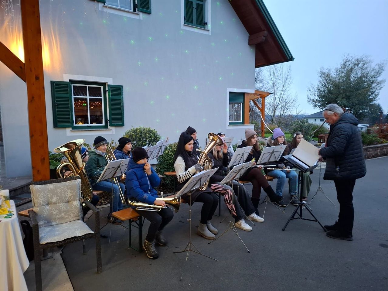 Eine Gruppe von Musikern spielt vor einem Haus. Sie tragen Winterkleidung und einige sitzen auf Stühlen. Ein Mann steht und spielt Trompete.