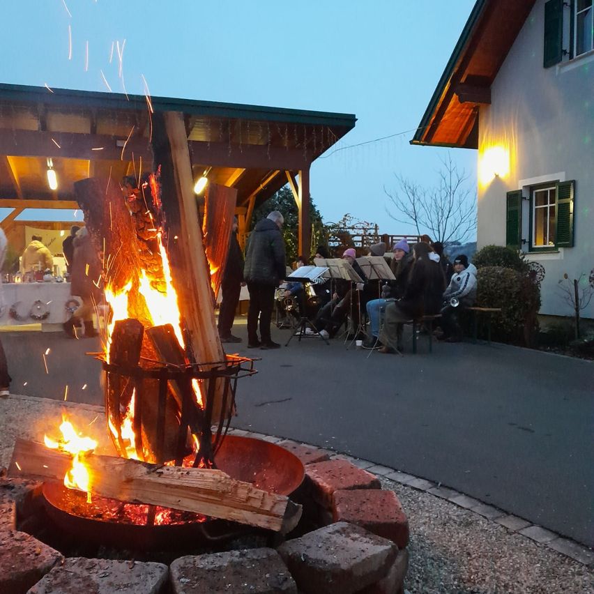 Ein Lagerfeuer brennt hell bei Dämmerung, umgeben von Menschen und Musikinstrumenten, mit einem Haus und Bäumen im Hintergrund.