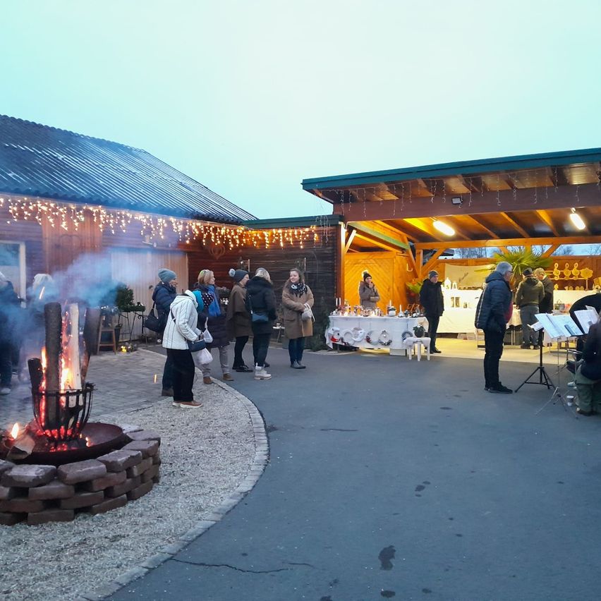 Eine Gruppe von Menschen steht um ein Feuerbecken in einem Außenbereich bei Nacht. Sie tragen Winterkleidung. Ein Tisch mit verschiedenen Gegenständen steht unter einem Vordach.