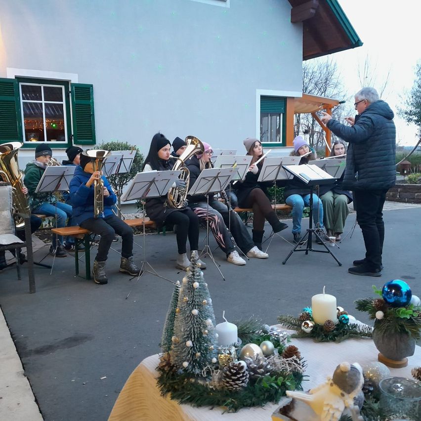 Eine Gruppe junger Musiker tritt draußen in einem festlichen Rahmen auf. Sie sitzen auf Bänken mit Notenständern vor ihnen. Ein Mann in einem schwarzen Jackett dirigiert sie.