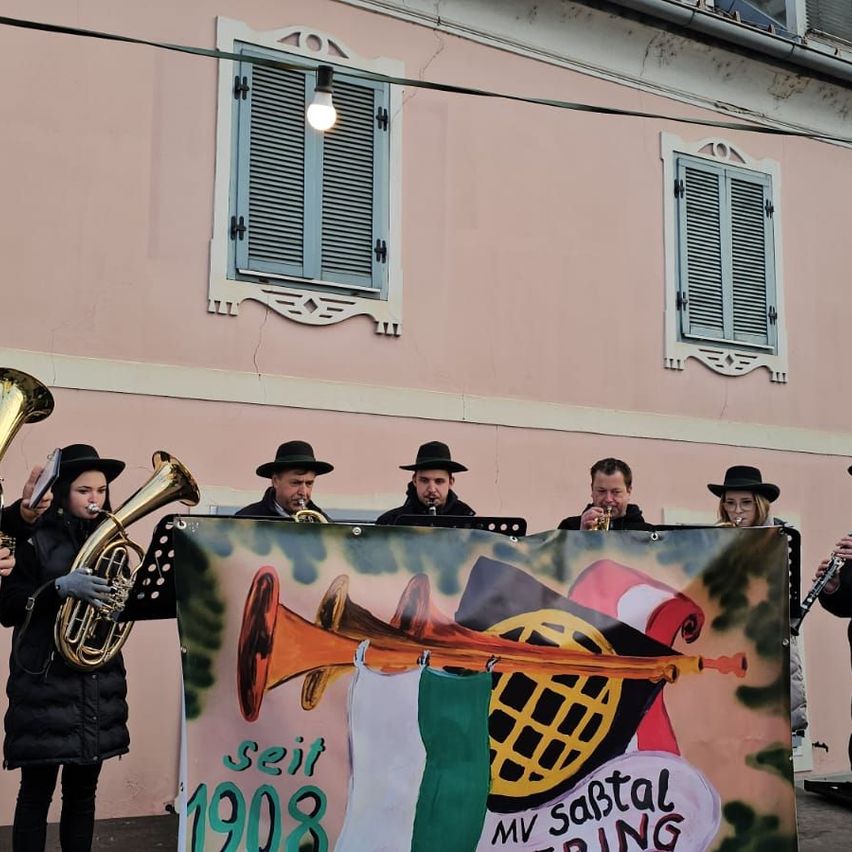 Eine Gruppe von Musikern in schwarzen Hüten und Mänteln tritt auf einer Bühne auf, vor ihnen ein Banner. Das Banner trägt den Text 'seit 1908 MV Sabtal Spring' und eine Illustration von Musikinstrumenten. Das Gebäude hinter ihnen hat rosa Wände und weiße Fensterläden.