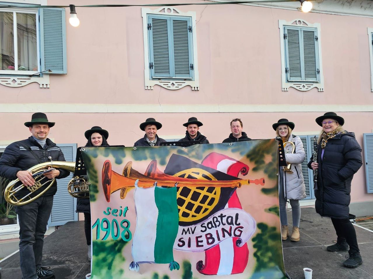 Sieben Musiker stehen vor einem Banner, das Musikinstrumente, eine Flagge und den Text 'MV Samstag Siebing' zeigt. Das Gebäude hinter ihnen hat drei Fenster mit Fensterläden.