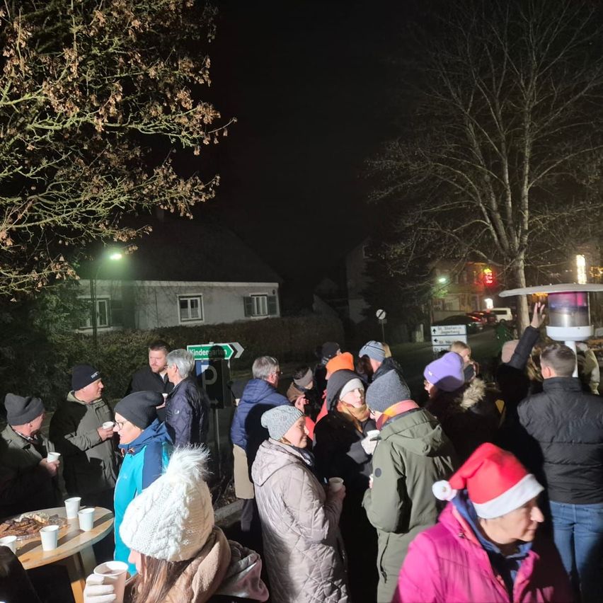 In der Nacht versammeln sich eine Gruppe von Menschen im Freien, einige in Winterkleidung und Mützen. Sie scheinen in Gespräche vertieft zu sein, möglicherweise bei einer gesellschaftlichen Veranstaltung. Im Hintergrund befindet sich ein Haus, Straßenlaternen und Bäume.