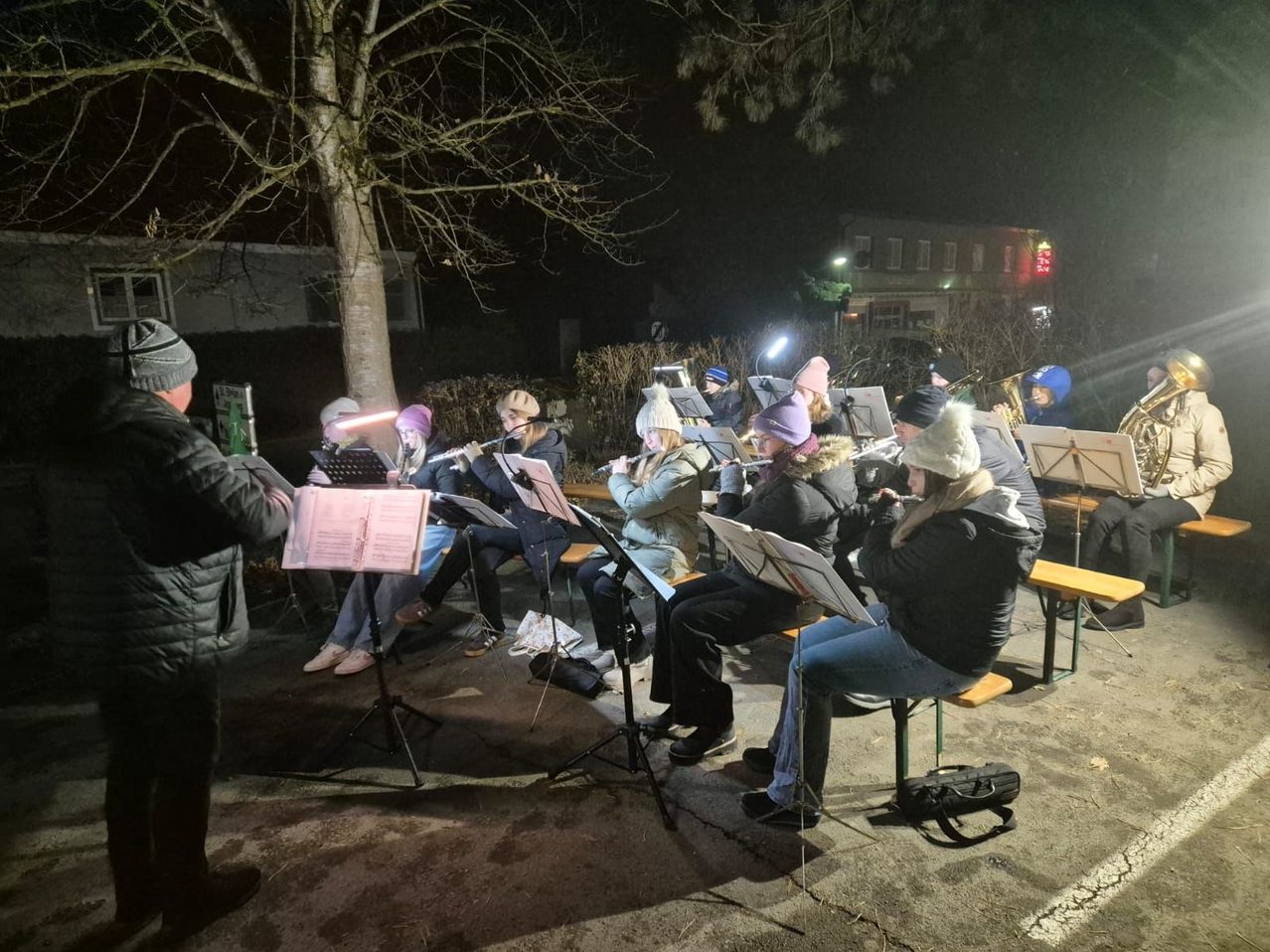 Eine Gruppe von Menschen sitzt bei Nacht draußen auf Bänken und spielt Musikinstrumente. Sie sind warm gekleidet, und ein Scheinwerfer beleuchtet sie. Die Umgebung ist vor einem Gebäude mit beleuchteten Fenstern.