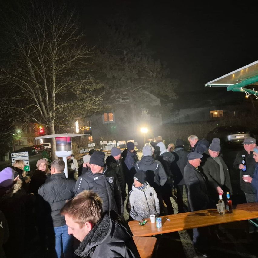 In der Nacht versammeln sich eine Gruppe von Menschen in Winterkleidung um einen Tisch im Freien, wahrscheinlich für eine Veranstaltung. Ein beleuchteter Heizstrahler und Flaschen stehen auf dem Tisch.