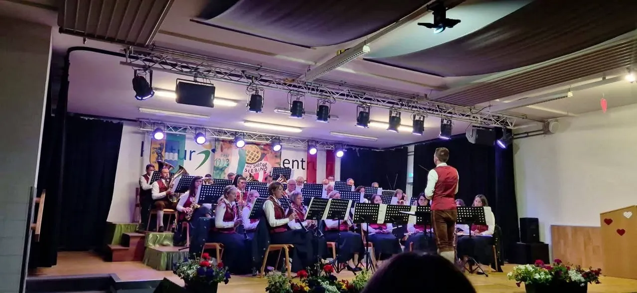 Ein Konzert auf der Bühne mit Musikern, die verschiedene Instrumente spielen, darunter Klarinetten und Trompeten. Die Bühne ist mit Blumen und Bannern geschmückt. Ein Scheinwerfer konzentriert sich auf einen Dirigenten vor der Bühne.