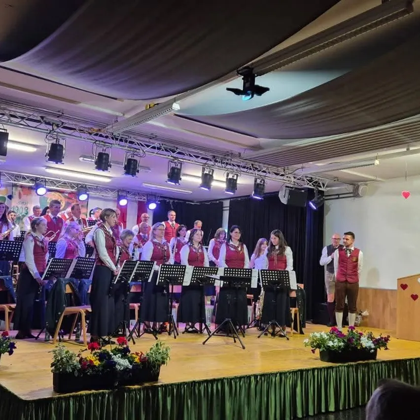 Ein Chor tritt mit Musikinstrumenten auf der Bühne auf, im Hintergrund eine Flagge. Die Bühne ist mit Blumen und Scheinwerfern geschmückt.