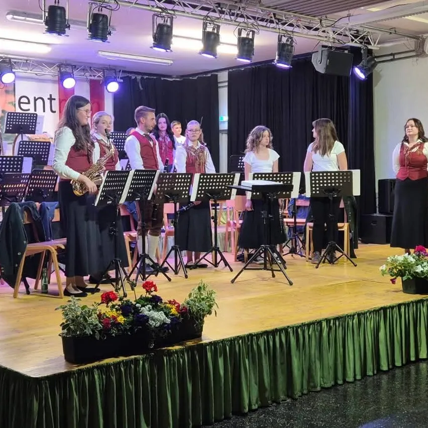 Eine Gruppe von Künstlern auf der Bühne mit Notenständern, Blumen und Bühnenlampen. Eine Frau spielt Saxophon.