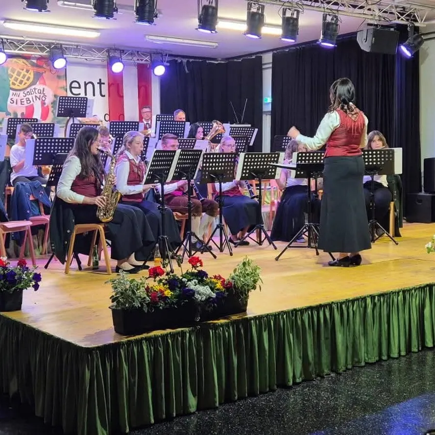 Eine Band tritt auf einer mit Blumen und grünen Vorhängen geschmückten Bühne auf. Eine Frau dirigiert die Musiker, während andere Saxophon, Trompete und Posaune spielen.