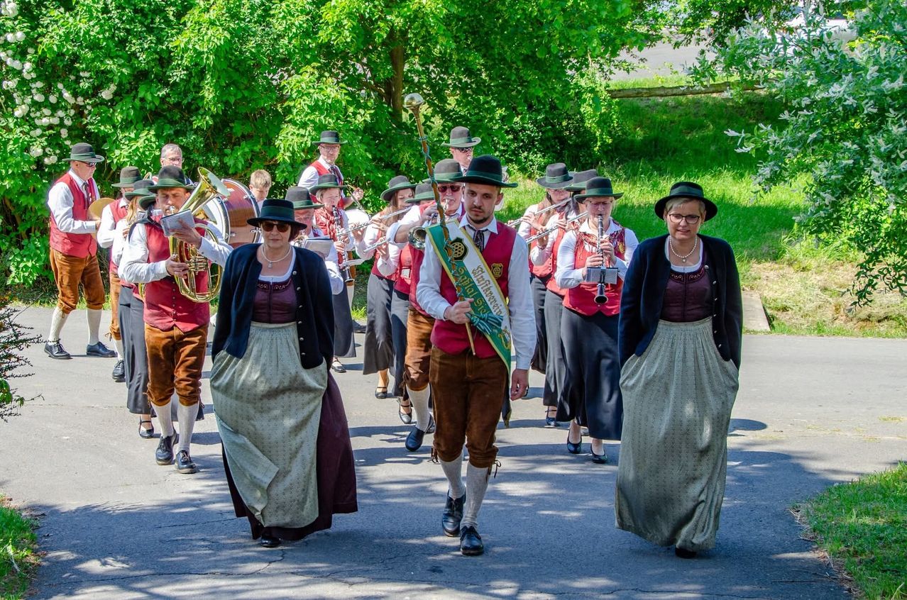 A marching band in traditional attire walks down a road, accompanied by musicians playing various instruments. The group is led by a man with a sash.