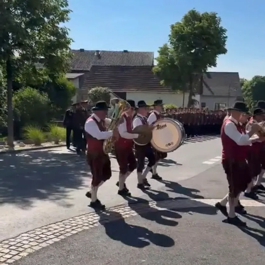 Eine Blaskapelle spielt auf der Straße und trägt traditionelle Kleidung. Sie spielen verschiedene Instrumente, darunter Tuben und Trommeln. Die Umgebung ist im Freien, mit Gebäuden und Bäumen im Hintergrund.