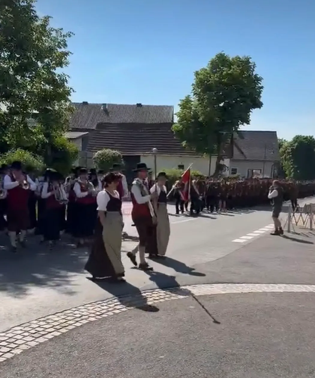 Eine Gruppe von Menschen in traditioneller Kleidung mit Hüten geht auf einer Straße, begleitet von einer Flagge und Zuschauern. Sie sind von Häusern und Grünflächen umgeben.