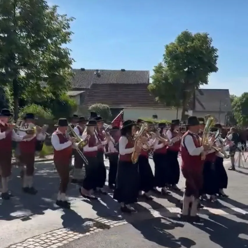 Eine Blaskapelle in traditioneller Kleidung marschiert auf einer Straße mit Gebäuden und Bäumen im Hintergrund.