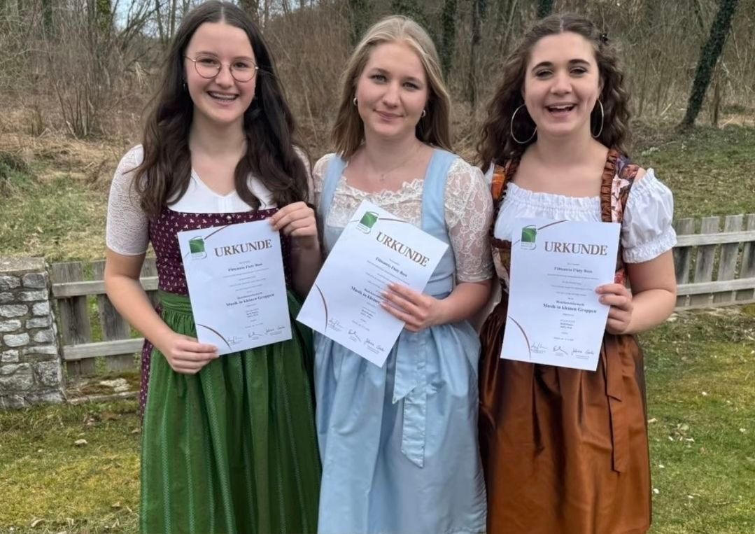Drei Frauen stehen in einem Waldgebiet, lächeln und halten Urkunden in den Händen. Sie tragen traditionelle deutsche Kleider. Die Frau auf der linken Seite trägt eine Brille. Die Frau in der Mitte trägt eine Halskette. Die Frau auf der rechten Seite trägt Ohrringe. Die Urkunden haben das Wort 'URKUNDE' darauf.
