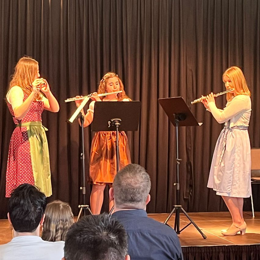 Drei junge Frauen spielen Flöten auf einer Bühne mit schwarzem Vorhang im Hintergrund. Die Frau links trägt ein gepunktetes Kleid, die anderen zwei Kleider. Mehrere Menschen sehen vom Publikum zu.