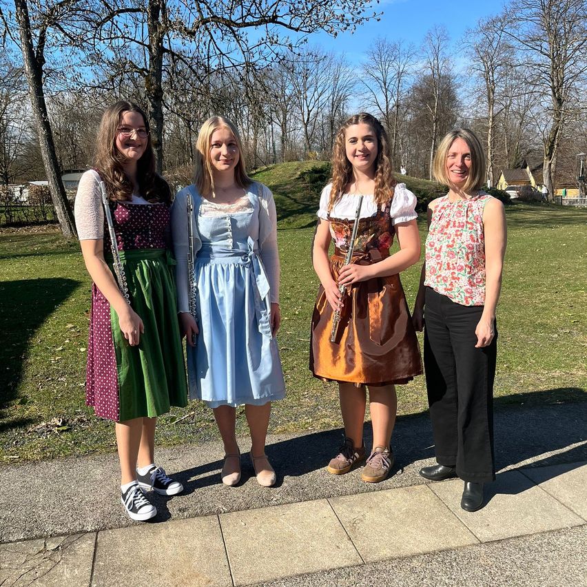 Vier Frauen stehen in einem Park. Drei von ihnen tragen traditionelle Kleider und halten Flöten. Die vierte Frau trägt ein Blumenoberteil und schwarze Hose. Hinter ihnen befindet sich eine Rasenfläche mit Bäumen.