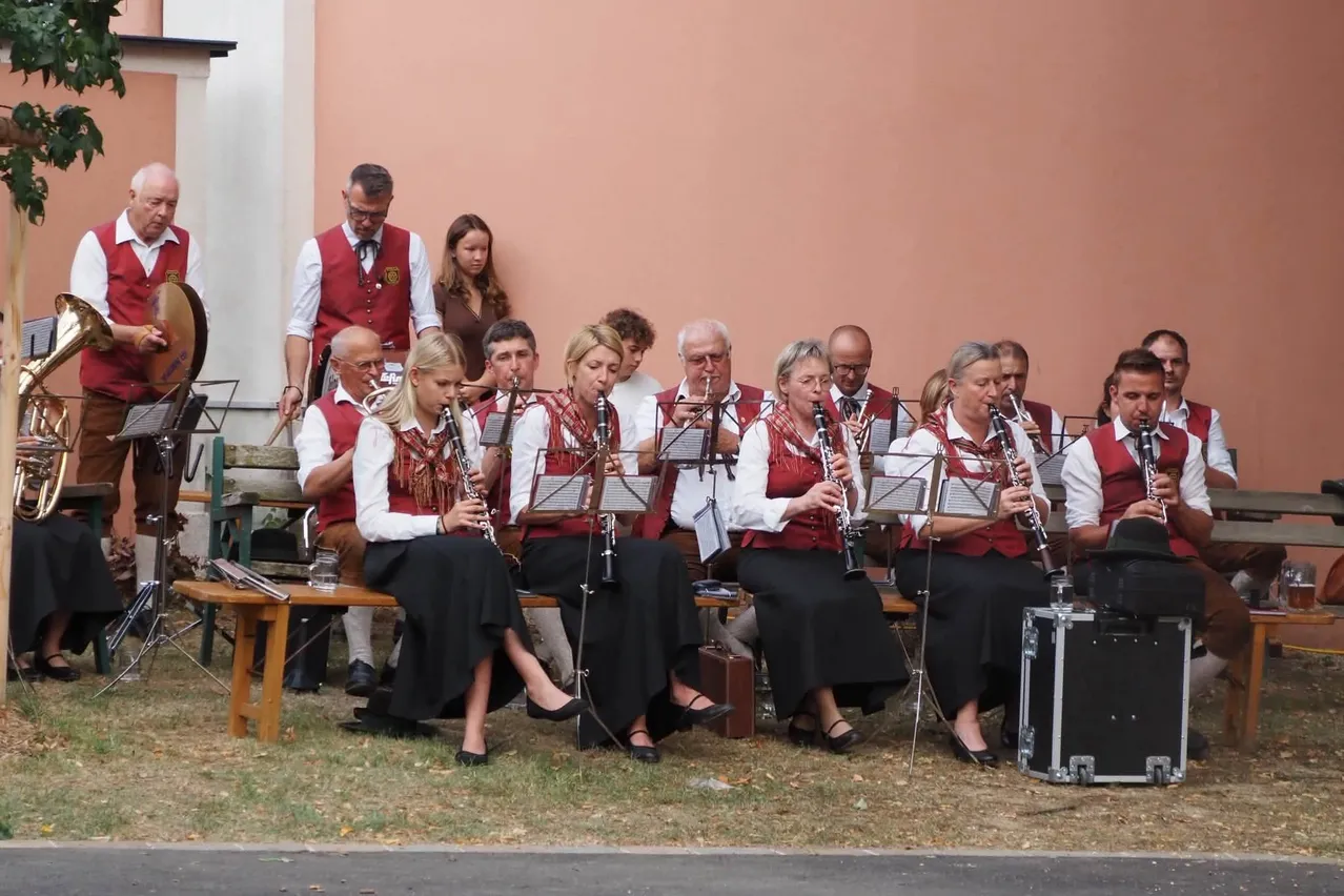 Eine Gruppe von Musikern in traditioneller Kleidung, die Klarinetten und Trompeten spielen, sitzt auf einer Bank vor einem Gebäude.