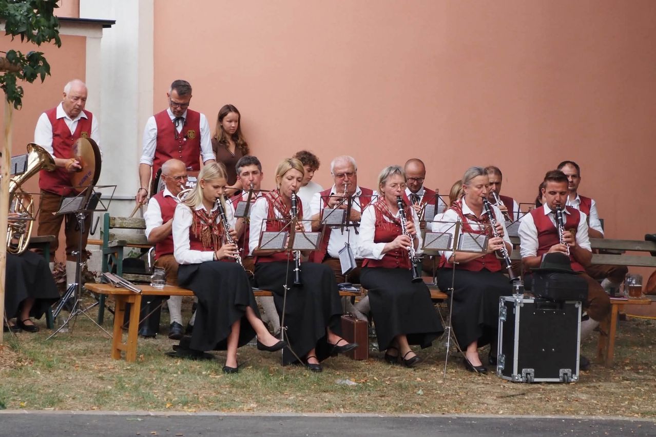A group of musicians in traditional attire, playing clarinets and trumpets, seated on a bench in front of a building.