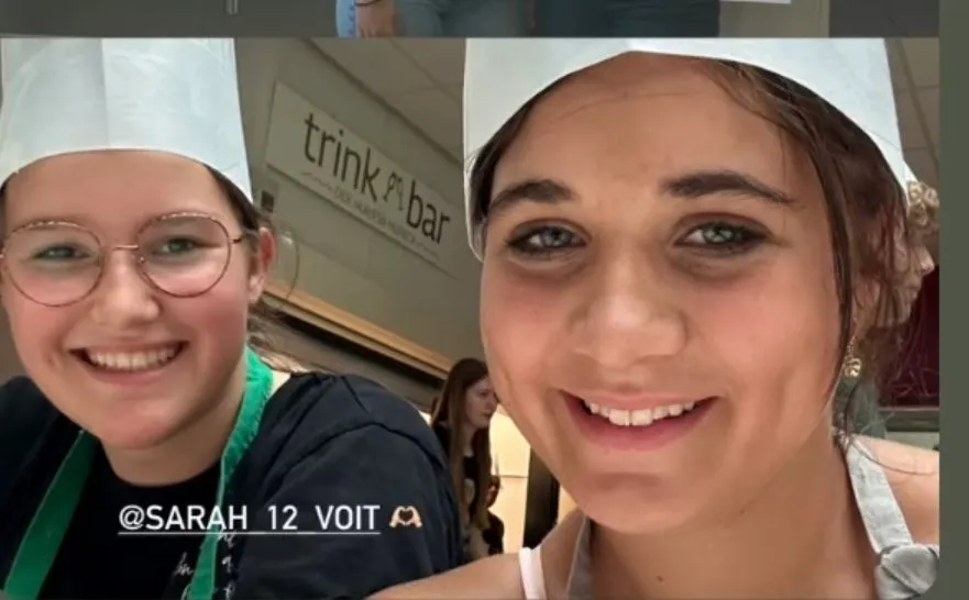 Zwei Frauen in Kochmützen lächeln für ein Foto vor einem Schild, das 'trink & bar' steht. Die Frau auf der linken Seite trägt eine Brille und eine grüne Lanyard.