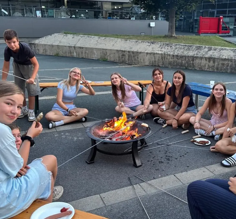 Eine Gruppe junger Leute sitzt um ein Lagerfeuer herum und röstet Marshmallows an Stöcken. Sie sitzen auf Bänken und am Boden, eine Person steht in der Nähe. Die Szene spielt im Freien, mit einer Betonbarriere und einem Gebäude im Hintergrund.