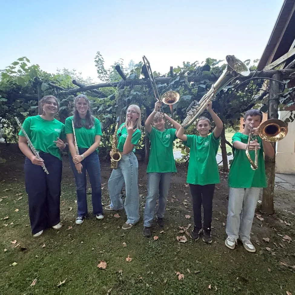 Eine Gruppe junger Musiker in grünen T-Shirts posiert mit ihren Instrumenten im Garten. Sie halten eine Posaune, Trompete und Flöte.