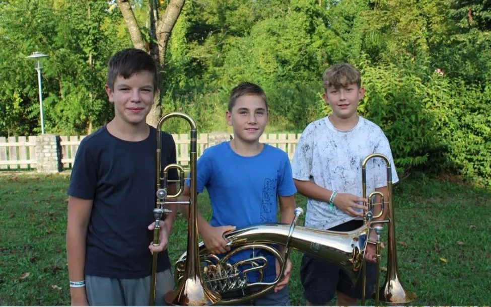 Drei Jungen stehen in einem Grasbereich, jeder hält ein Blechblasinstrument. Der Junge auf der linken Seite hält ein Tenorhorn, der Junge in der Mitte hält eine Tuba und der Junge auf der rechten Seite hält ein Waldhorn. Sie lächeln und scheinen für ein Foto zu posieren.