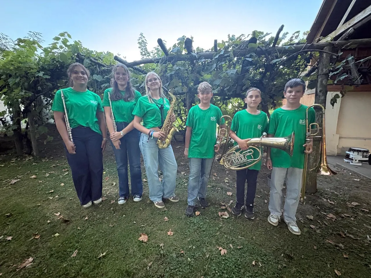 Eine Gruppe junger Musiker in grünen T-Shirts posiert für ein Foto, jeder hält ein anderes Instrument. Sie stehen auf einer Rasenfläche mit einer Holzstruktur und Grünpflanzen im Hintergrund.