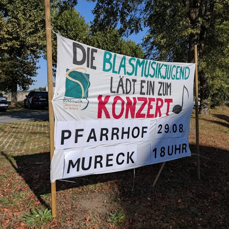 Ein weißes Banner mit grünem und rotem Text lautet 'Die Blasmusikjugend lädt ein zum Konzert Pfarrhof Mureck 29.08. 18UHR'. Das Banner ist auf einem Grasbereich mit Bäumen und geparkten Autos im Hintergrund angebracht.