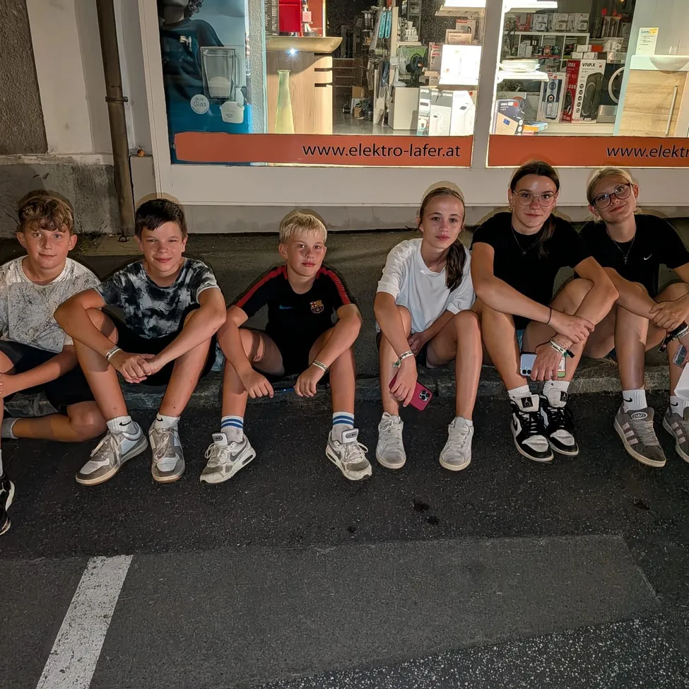 Fünf Teenager sitzen auf einem Bordstein vor einem Geschäft mit einem Glasfenster, das verschiedene Produkte anzeigt.